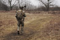 A soldier in camouflage walking along a grassy path through a forested area.